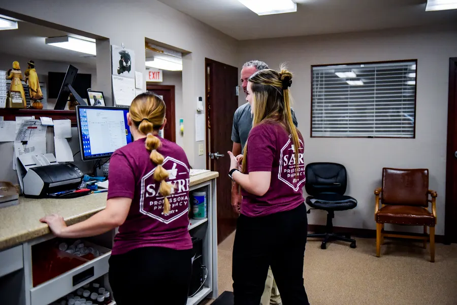 Two pharmacy team members working at computers behind the prescription counter.