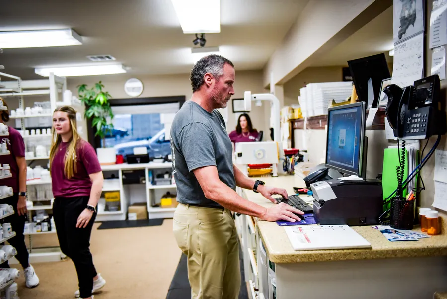 A pharmacist working at a computer behind the counter with pharmacy team members nearby.