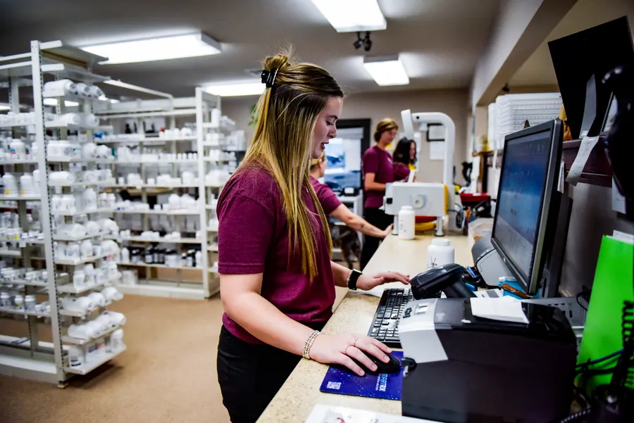 A pharmacy team member working at a computer beside shelves of prescription medicines.