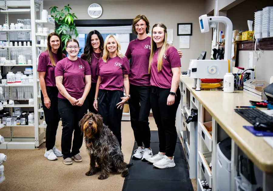 Six pharmacy team members gathered behind the counter with a dog seated in front.